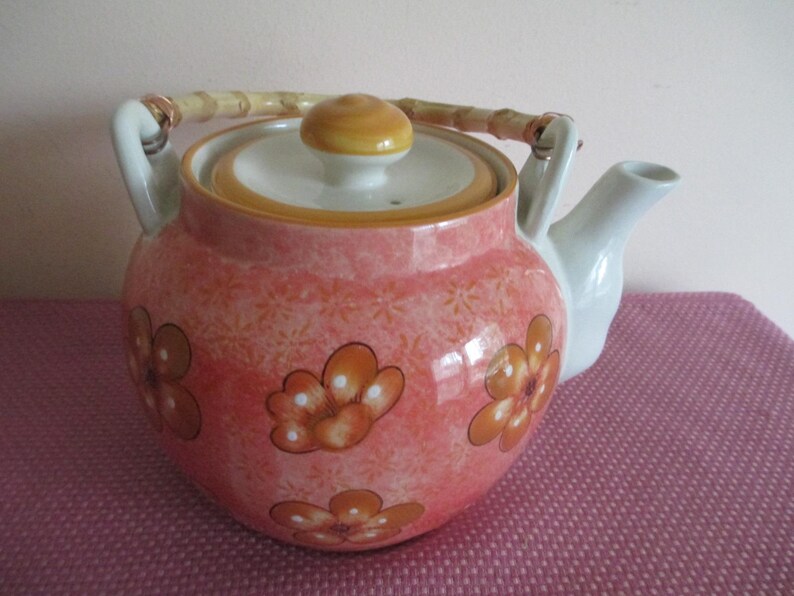 K&ouml;nnte beinhalten: Eine Keramik-Teekanne mit Bambusgriff und Deckel. Die Teekanne ist haupts&auml;chlich rosa mit einem Blumenmuster und orangefarbenen Blumendesigns. Der Ausguss und die Griffbefestigungen sind wei&szlig;.