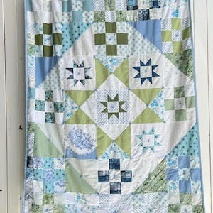 Peut inclure: Une courtepointe faite à la main avec un motif d'étoiles dans des tons de bleu, vert et blanc. La courtepointe présente divers motifs de tissu, y compris des motifs floraux et géométriques, et est bordée d'une bordure bleu clair.