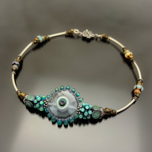 Może przedstawiać: Srebrny naszyjnik z łańcuszkiem z dużą, przezroczystą szklaną koralikiem z wzorem niebieskiego i zielonego oka. Koralik jest otoczony mniejszymi, turkusowymi i brązowymi szklanymi koralikami.