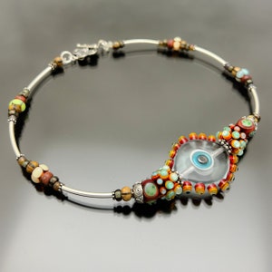 Może przedstawiać: Srebrna bransoletka z przezroczystą szklaną koralikiem w kształcie oka, otoczonym kolorowymi koralikami w odcieniach czerwieni, pomarańczy, żółci, zieleni i błękitu.