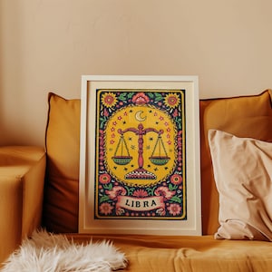 Op de afbeelding: Een ingelijste Libra dierenriemkunstprint. Het kunstwerk heeft een gele achtergrond met een weegschaalsymbool, omringd door bloemen- en sterontwerpen. Het woord "LIBRA" wordt op een banner onderaan weergegeven. De lijst is wit.