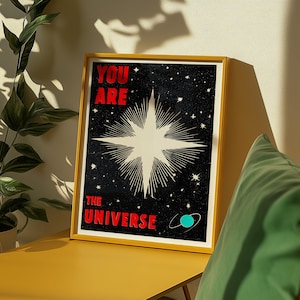 May include: A framed poster with a yellow border. The poster features the words "YOU ARE THE UNIVERSE" in red, with a large starburst design in the centre against a black background. Small white stars and a teal planet graphic are also visible.