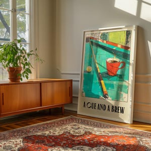 May include: A framed print of a green pool table with a cue stick, a white billiard ball, and a red coffee mug with steam rising from it. The text "A CUE AND A BREW" is printed below the image.