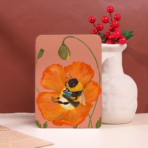 Op de afbeelding: Een perzikkleurige telefoonhoes met een illustratie van een hommel die een boek leest op een oranje klaproos. De bij is zwart en geel, en de bloem heeft groene stelen en bladeren. Een witte vaas met rode bessen staat op de achtergrond.