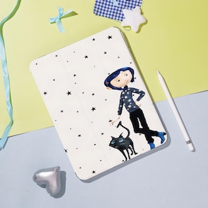 Pode incluir: Capa de tablet branca com uma ilustração de desenho animado de uma menina, um gato preto e estrelas pretas. A menina usa um suéter azul com estrelas brancas, calças pretas e botas azuis. Uma caneta branca também está presente.