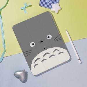 Può includere: Custodia grigia per tablet con un volto di personaggio dei cartoni animati, grandi occhi e baffi. La custodia ha una sezione inferiore bianca. Uno stilo bianco è accanto alla custodia. Lo sfondo è azzurro e giallo.