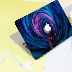 May include: Laptop with a custom cover featuring a whimsical illustration. The design showcases a figure with an Apple logo head, dressed in a red jacket, set against a swirling blue and purple tunnel background. The laptop is on a yellow and white surface.