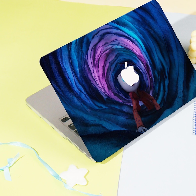 May include: Laptop with a custom cover featuring a whimsical illustration. The design showcases a figure with an Apple logo head, dressed in a red jacket, set against a swirling blue and purple tunnel background. The laptop is on a yellow and white surface.