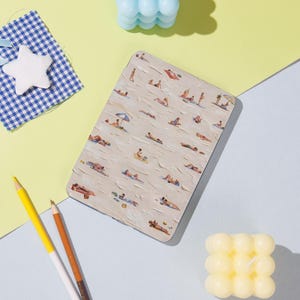 May include: Rectangular notebook with a beach scene illustration. The cover shows a beige background with figures sunbathing, some under umbrellas. The notebook is surrounded by yellow and blue objects, including pencils and candles.