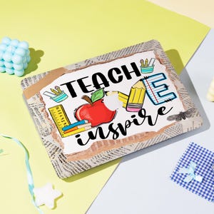 Può includere: Un cartello rettangolare con le parole "TEACH inspire" in nero. Il design include una mela rossa, matite, un righello e un quaderno. Il cartello è incorniciato con carta di giornale.