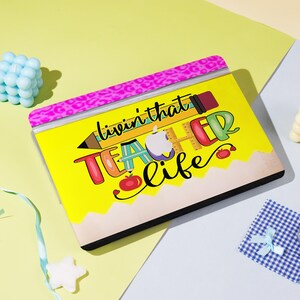 Può includere: Una custodia gialla per laptop con un design a matita e le parole "livin' that TEACHER life" in lettere colorate. La custodia ha un bordo rosa con stampa leopardata e un bordo inferiore a forma di matita. Il design include una mela, un righello e mele rosse.