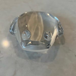 Puede incluir: Una figurita de rana de vidrio transparente con una superficie lisa y redondeada. La rana tiene dos ojos grandes y redondos y una boca ligeramente abierta.