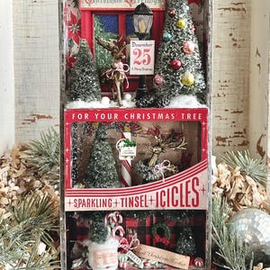 Peut inclure: Une boîte d'ombre sur le thème de Noël avec des arbres miniatures, des rennes et des décorations festives. La boîte présente le texte "Christmas Greetings" et "December 25". La section inférieure indique "Sparkling + Tinsel + Icicles".