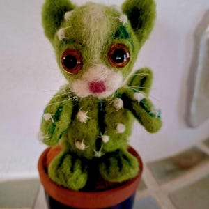 Op de afbeelding: Een naaldgevilte groene cactusfiguur met grote bruine ogen, een witte snuit en een roze neus. Het figuurtje heeft kleine witte stekels en staat in een kleine terracotta-kleurige pot met een donkerblauwe onderkant.