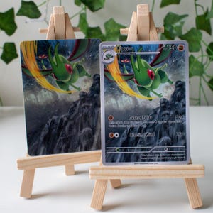 Puede incluir: Dos cartas de juego de cartas coleccionables con el personaje Flygon, exhibidas en pequeños caballetes de madera. Las cartas tienen una combinación de colores verde y amarillo con el texto "Flygon" y otros detalles relacionados con el juego.