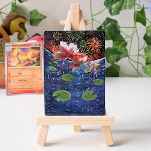 May include: A rectangular art print on a small wooden easel. The print depicts a koi fish swimming in a pond with lily pads, fireworks, and a dark blue background. The koi fish is red and white.