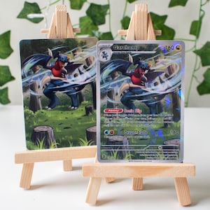 Puede incluir: Dos cartas coleccionables Pokémon Garchomp exhibidas en pequeños caballetes de madera. Las cartas presentan una criatura ilustrada azul, roja y blanca en un entorno forestal. Una carta tiene texto que incluye "Sonic Slip" y "Dragonblade".