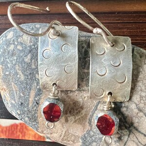 May include: A pair of silver earrings with rectangular, textured pendants featuring circular cutouts. Each earring has a faceted, deep red bead dangling below. The earrings are displayed on a gray stone surface.