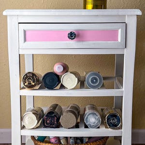 May include: White shelf unit with three shelves, a pink drawer, and a crystal knob. The shelves are filled with spray paint cans in various colors. A wicker basket sits on the bottom shelf, filled with smaller bottles of paint. A gold can with paintbrushes sits on top of the shelf unit.