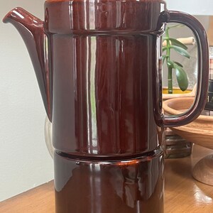 May include: A brown ceramic coffee pot with a lid and a handle. The pot has a base that is separate from the main body.