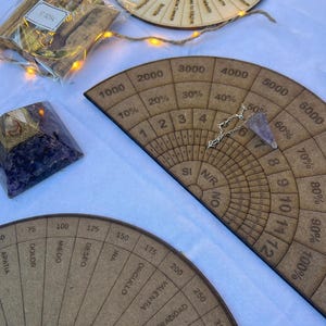 Puede incluir: Una colección de herramientas espirituales, que incluye péndulos de madera, una pirámide de cristal y un tablero de adivinación de madera con números y porcentajes. Los artículos están dispuestos sobre una superficie blanca.