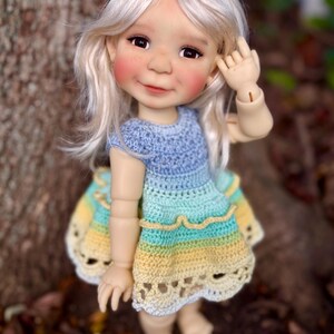 May include: A handcrafted doll with fair skin, rosy cheeks, and blonde hair. The doll wears a blue, yellow, and green crocheted dress. The doll is standing in front of a tree.