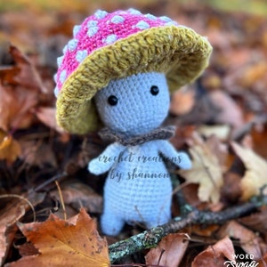 Könnte beinhalten: Eine gehäkelte Pilzfigur mit einer rosa-weißen Kappe, einem gelben Rand und einem grauen Körper. Die Figur steht auf einem Bett aus Herbstlaub. Der Text "crochet creations by shannon" ist auf dem Körper der Figur sichtbar.