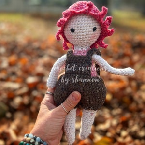 May include: A crocheted doll with a brown and white dress, pink hair, and black eyes. The doll is being held by a hand wearing bracelets.