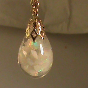 May include: A teardrop-shaped pendant with a gold-colored cap and chain. The clear glass pendant is filled with white, iridescent fragments. The gold cap has a decorative design. The pendant is hanging.