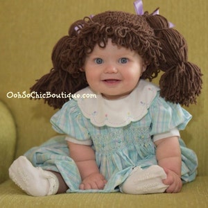 May include: A baby wearing a brown yarn doll wig with pigtails and a light blue and white plaid dress. The baby is sitting on a green chair. The website OohSoChicBoutique.com is visible in the image.