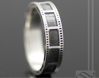 May include: A silver ring with a film strip design. The ring is made of metal and has a brushed finish.