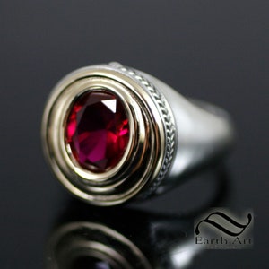 May include: A silver ring with a red gemstone set in a gold bezel. The ring has a braided design around the band.