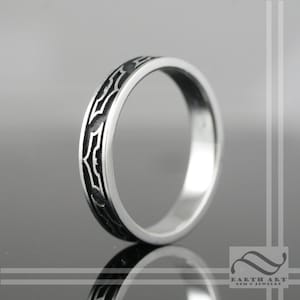 May include: A silver ring with a black engraved pattern. The ring has a thin band and a repeating design around the circumference. The ring is displayed on a reflective surface.