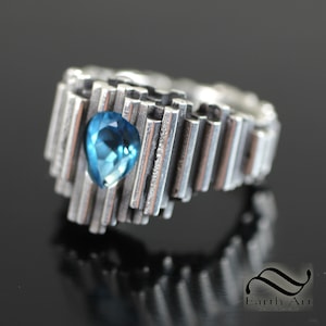 Puede incluir: Un anillo de plata con una piedra preciosa azul engastada en un diseño geométrico. El anillo está hecho de muchas pequeñas barras verticales.