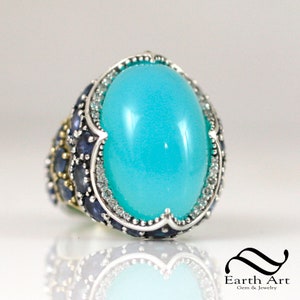 May include: A turquoise gemstone ring with a silver band and a halo of smaller gemstones. The ring is set with blue sapphires and diamonds.