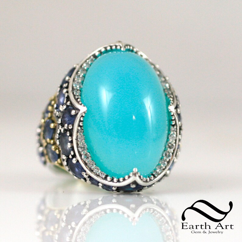 May include: A turquoise gemstone ring with a silver band and a halo of smaller gemstones. The ring is set with blue sapphires and diamonds.