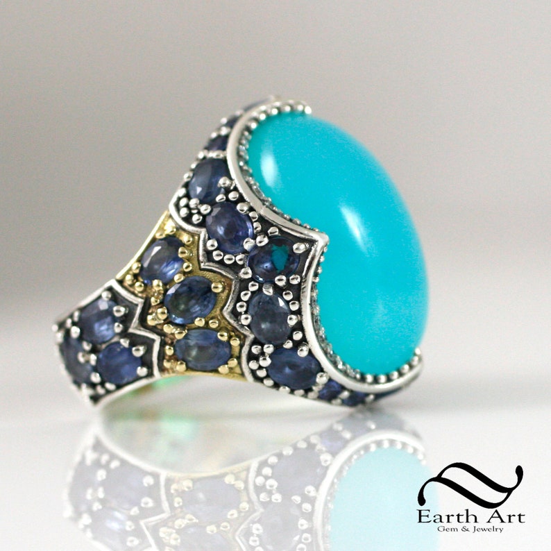 May include: A silver ring with a large turquoise cabochon stone set in a silver and gold setting. The setting is decorated with small blue sapphires.
