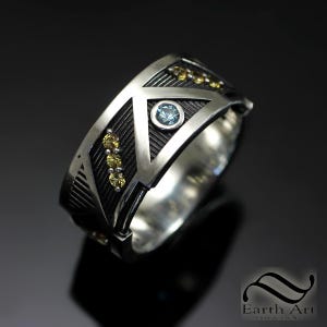 Könnte beinhalten: Ein silberner Ring mit geometrischem Design, der einen zentralen hellblauen Edelstein und kleinere gelbe Edelsteine aufweist. Der Ring hat einen strukturierten schwarzen Hintergrund innerhalb der geometrischen Formen, vor dunklem Hintergrund.