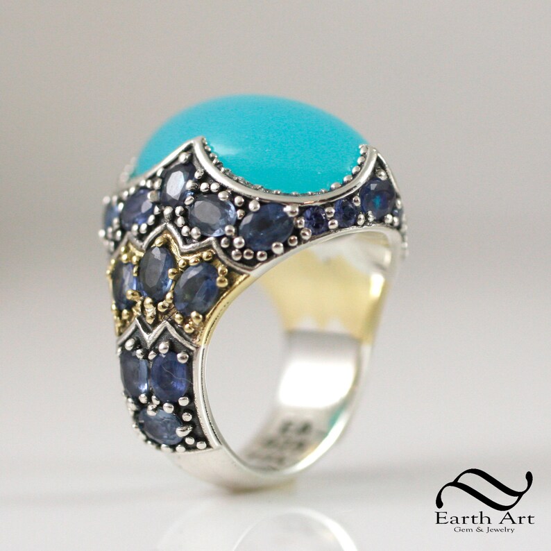 May include: A silver ring with a large turquoise cabochon stone set in a gold-toned bezel. The ring is decorated with smaller blue gemstones around the stone and the band.