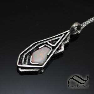 May include: A silver pendant necklace with an abstract design. The pendant features a central, light-colored opal stone, accented by black and red stones. The necklace has a textured silver chain and the pendant is approximately 2 inches long.