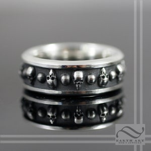 May include: A silver ring with a band of skulls and small silver balls. The ring is polished and has a high shine.