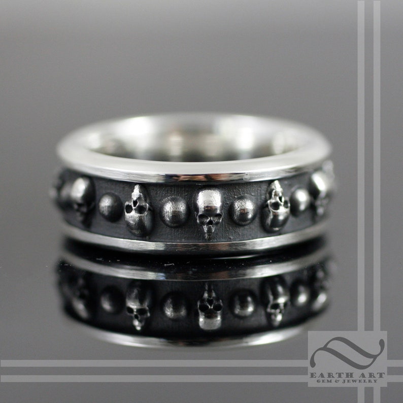 May include: A silver ring with a band of skulls and small silver balls. The ring is polished and has a high shine.