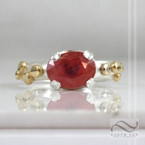 May include: A silver ring with a red gemstone set in a gold bezel. The ring is decorated with small gold beads.