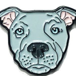 May include: A blue and black enamel pin featuring a cartoon pit bull dog with pink ears. The dog has a serious expression and blue eyes.