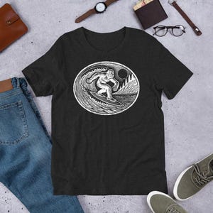 May include: A black t-shirt with a white graphic of a Bigfoot surfing on a wave. The graphic is in a circle with a moon and mountains in the background.