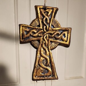 May include: A bronze-colored Celtic cross wind chime with intricate knotwork and three smaller crosses hanging below. The cross has a textured surface and is suspended by thin wires with black beads.