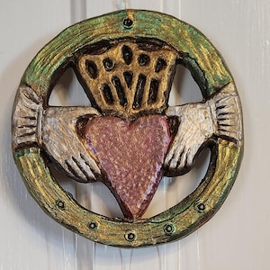May include: A Celtic knot wind chime with a green, gold, and silver design. The knot features a heart in the center and two hands clasped together. The chime has five hanging pieces with a brown and gold design.
