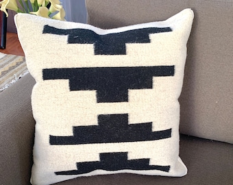 May include: A white throw pillow with a black geometric pattern. The pillow is on a brown couch with a white throw blanket.