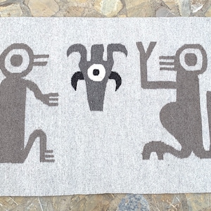 May include: A grey and white woven rug with a geometric design featuring two stylised figures and a central motif. The figures have large eyes and are kneeling with their arms raised.