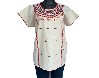 May include: A white cotton blouse with red embroidery around the neckline and down the sides. The embroidery features small red flowers and a geometric pattern.